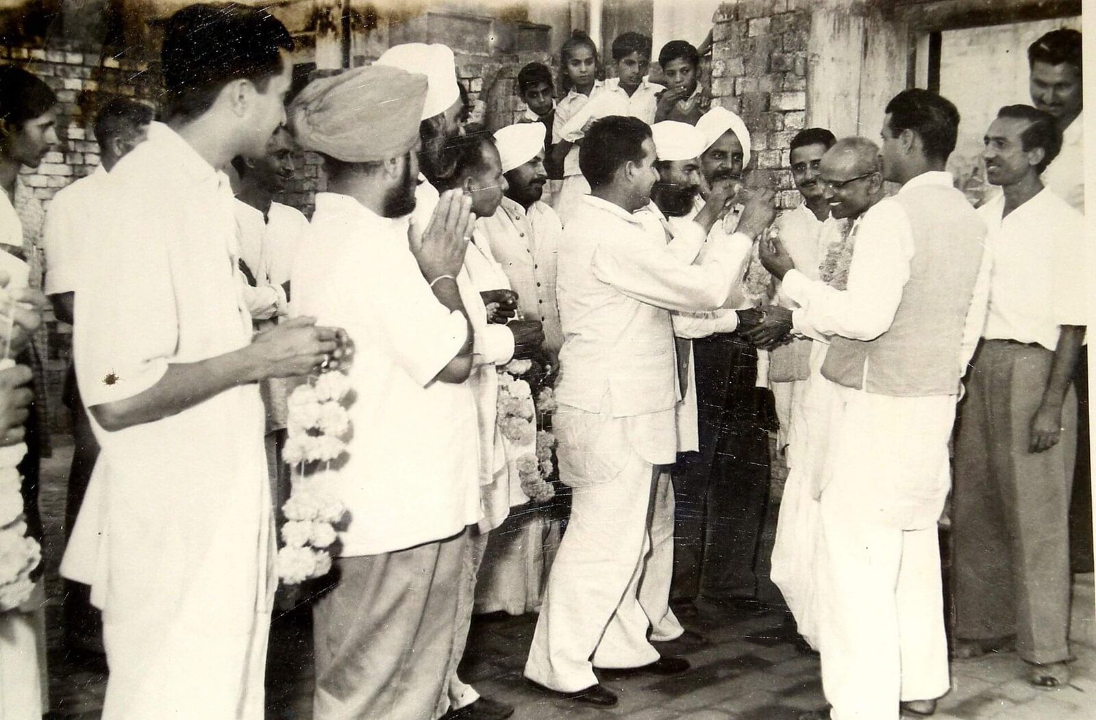 A large crowd of men in white clothing gathered outdoors beside a brick wall, garlanding Batukeshwar Dutt, who stands at the right with folded hands.